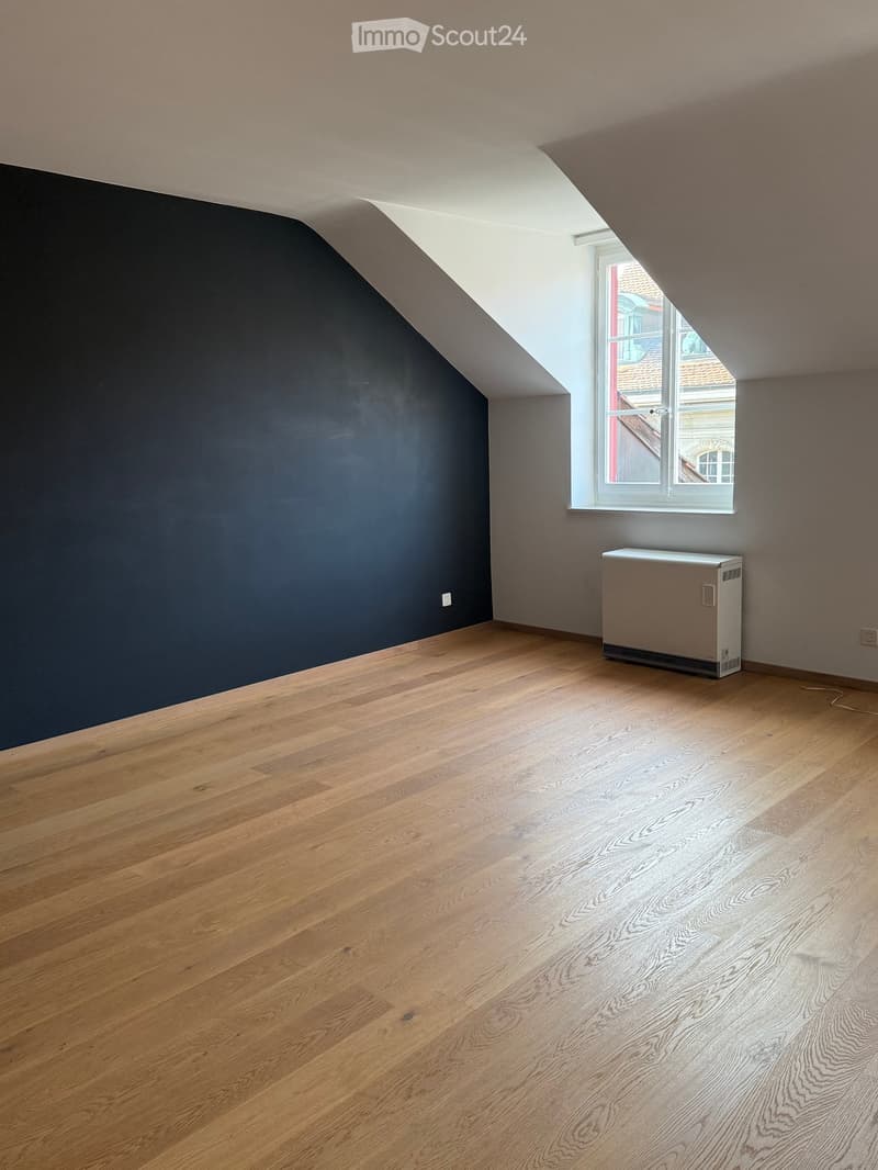 Empty attic room with wooden flooring, dark grey walls, and a single window letting in natural light. A radiator is visible against the wall.