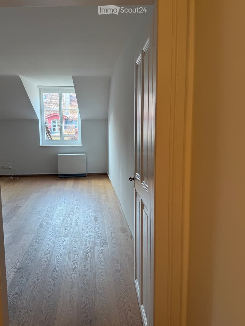 Empty room with wooden flooring, white walls, a window with a view of a roof, and a radiator. No furniture or equipment present.