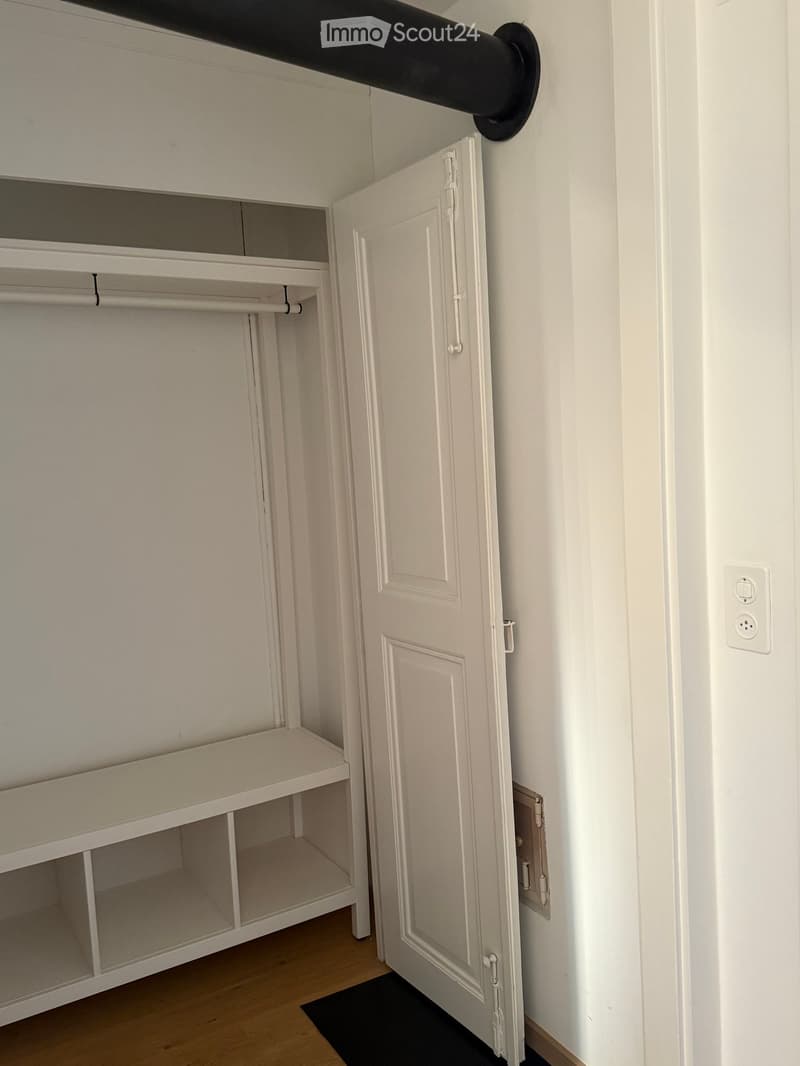 Empty hallway with white walls, wooden floor, open closet with empty shelves, and a closed door.