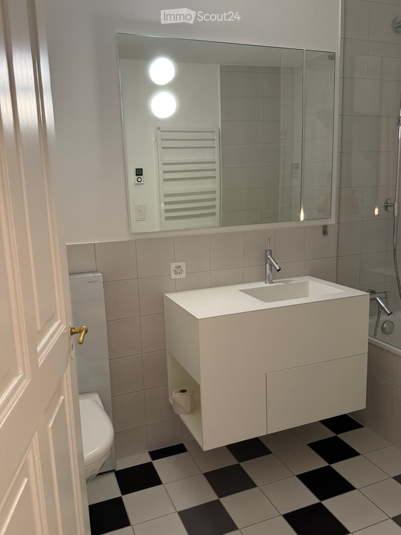 Modern bathroom with a white sink, chrome faucet, large mirror with built-in lights, toilet, radiator, and tiled floor in a black and white checkered pattern.