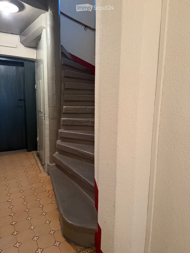A narrow hallway with a staircase leading upwards, featuring patterned tiles on the floor and a dark door on the left side.