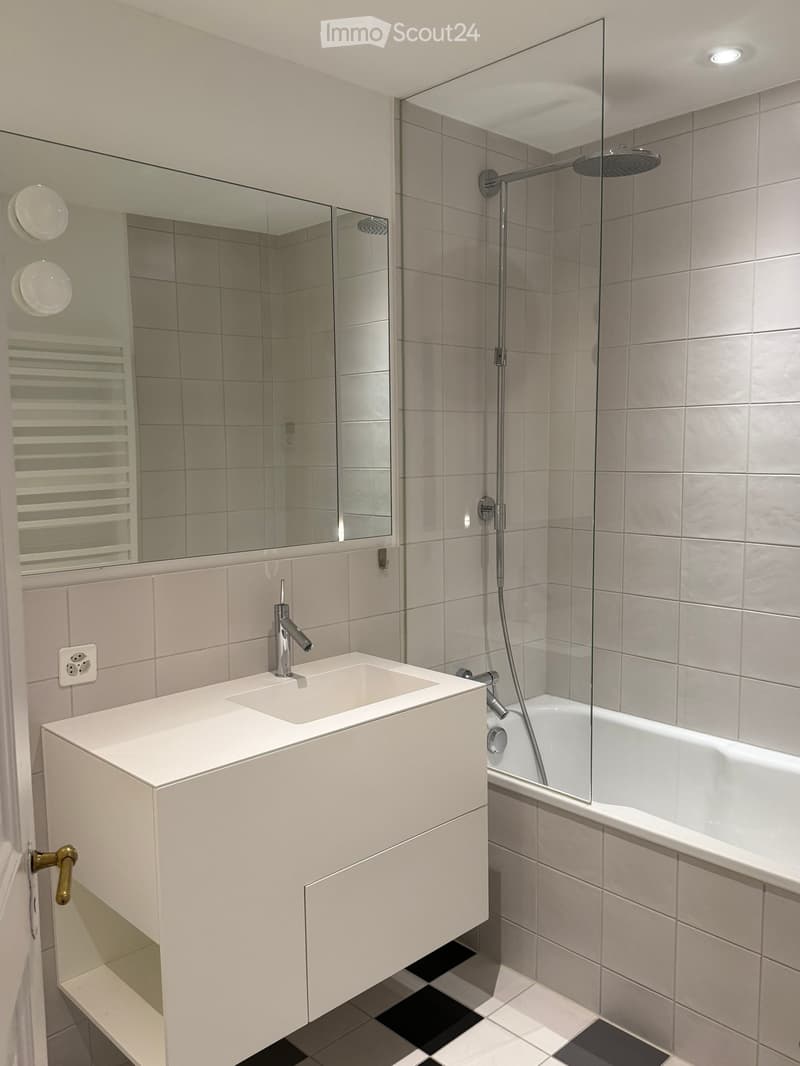 Modern bathroom with a white sink, chrome faucet, bathtub with glass shower enclosure, and a large mirror above the sink.