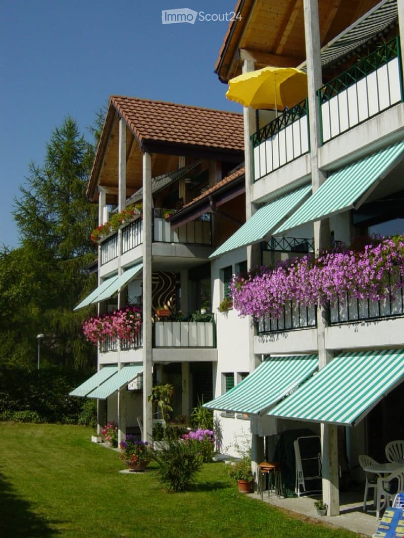 Multi-story residential building with balconies, green awnings, and flower boxes, surrounded by a well-maintained lawn and outdoor seating area.