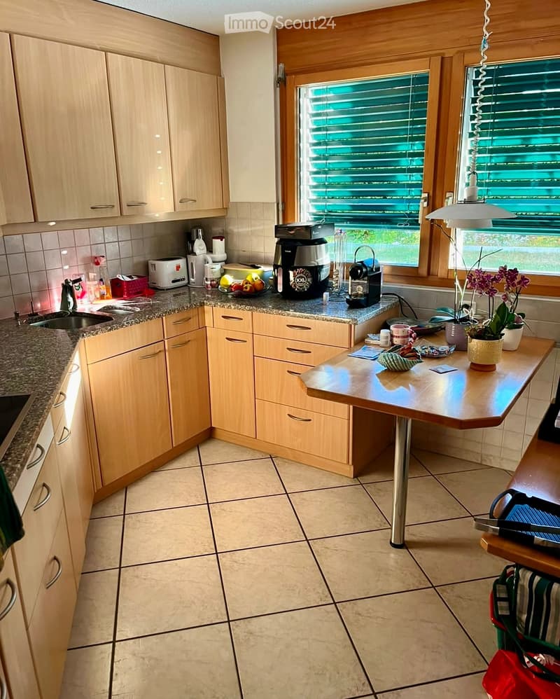 Bright kitchen with light wooden cabinets, granite countertops, and tiled floor. Features a coffee maker, toaster, sink, and a small table with a flower pot.