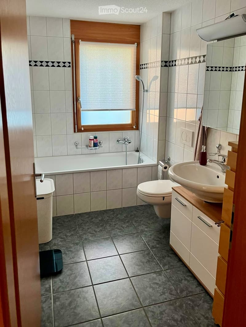 Modern bathroom with a bathtub, toilet, sink, mirror, and wooden cabinet. Features tiled walls and floor, a window with blinds, and a showerhead above the bathtub.