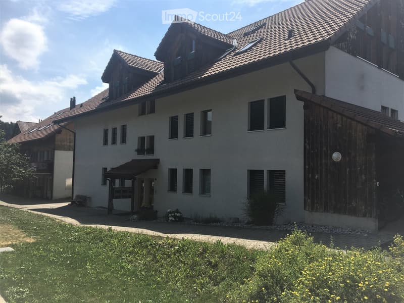 Two-story house with a tiled roof, white exterior walls, multiple windows, and a wooden garage. The house is surrounded by a well-maintained garden with flowers and a paved pathway leading to the entrance.