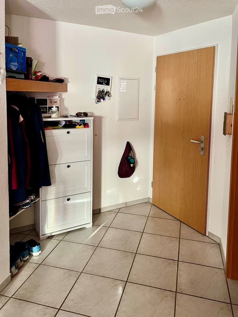 Hallway with tiled floor, white walls, coat rack, shoe rack, and open wooden door leading to another room.