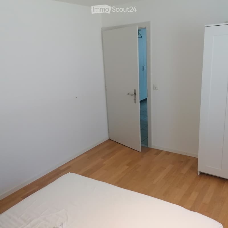 Bedroom with wooden flooring, white walls, a single bed, and a white wardrobe. An open door leads to another room.