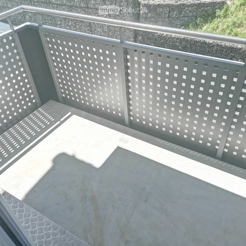 Modern balcony with grey patterned metal railings, concrete floor, and a stone wall in the background.