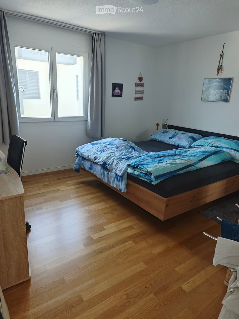 Bright bedroom with wooden flooring, a double bed with blue bedding, a chair, a small table, and a window with curtains.