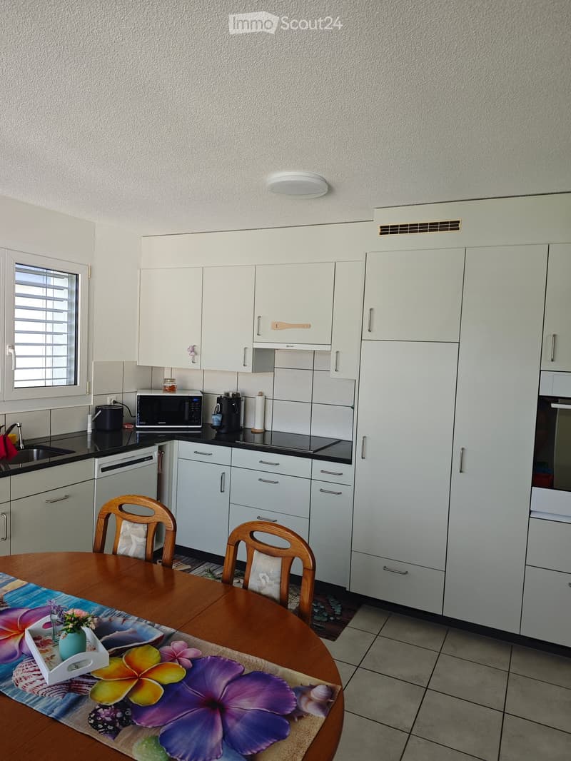 Modern kitchen with white cabinets, stainless steel appliances, and a wooden dining table with chairs. The table is set with a floral table runner and a small vase of flowers.