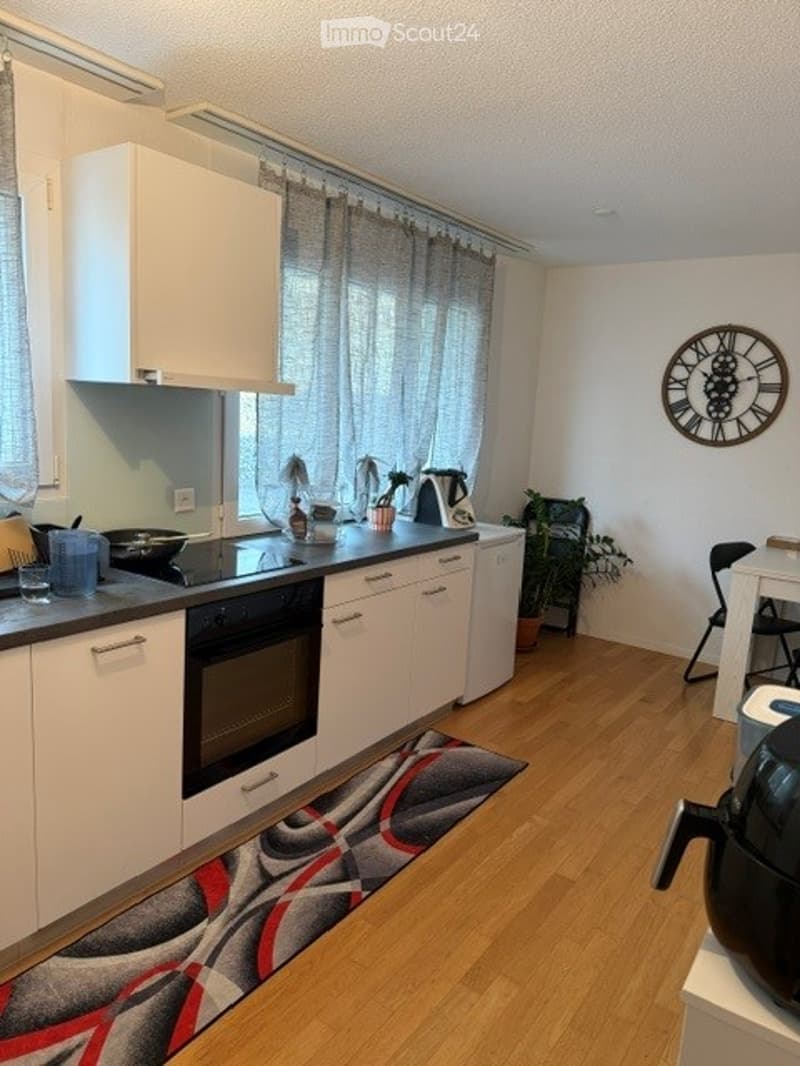 Modern kitchen with white cabinets, black oven, countertop, sink, and various kitchen items. Wooden flooring, a patterned rug, and a clock on the wall.