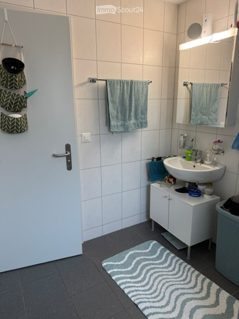 Modern bathroom with white tiled walls, a white sink, a mirror, a small cabinet, and a blue towel hanging on a rack. A decorative rug is placed on the floor.