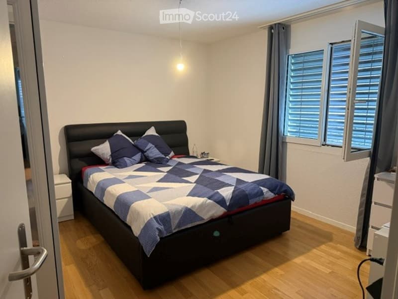 Modern bedroom with a large bed, geometric patterned blanket, pillows, and a nightstand. The room features wooden flooring and large windows with blinds.