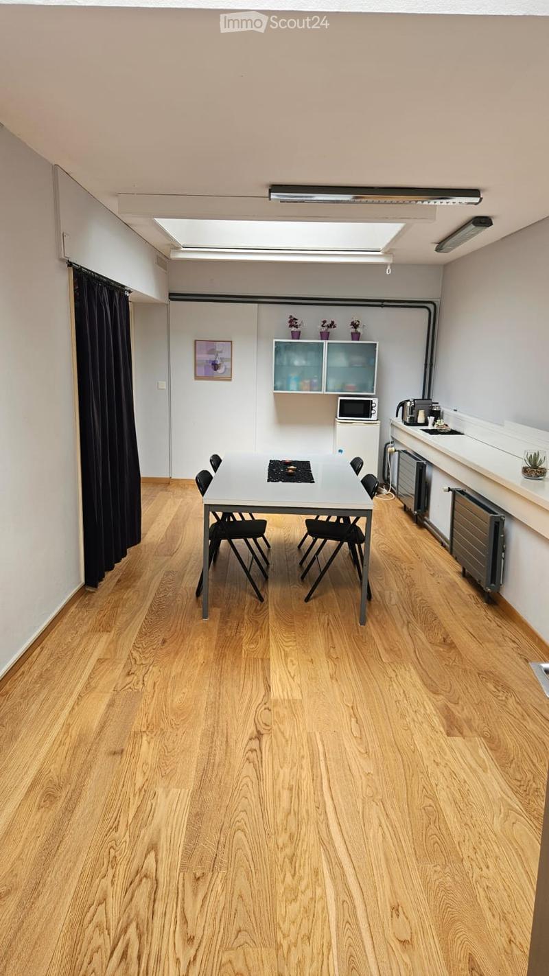A modern, well-lit room with wooden flooring, a dining table with chairs, a small kitchen area with a microwave, fridge, and radiator, and a picture frame on the wall.