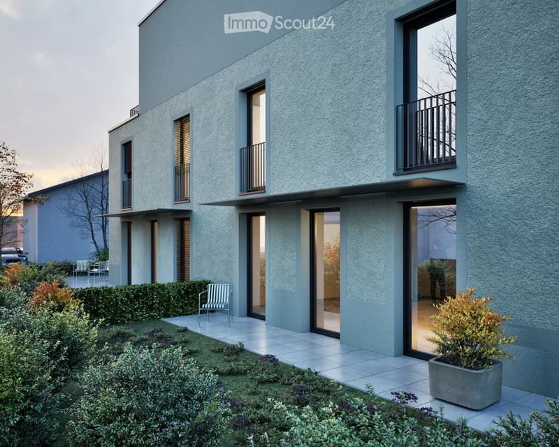 Modern two-story house with grey exterior, large windows, balconies, and a well-maintained garden with shrubs and potted plants.