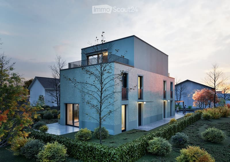 Modern two-story house with a combination of light blue and beige exterior walls, large windows, and a balcony on the upper floor. The house is surrounded by a well-maintained garden with shrubs and trees.
