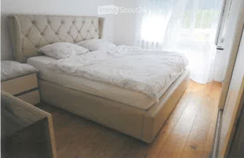 A well-lit bedroom with a large, upholstered bed, white bedding, and a matching nightstand with a mirror. The room features wooden flooring and sheer white curtains.