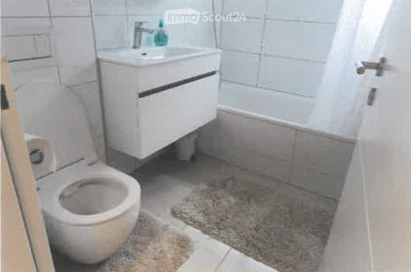 Modern bathroom with a white toilet, a sink with a chrome faucet, and a small bathroom cabinet. The floor is tiled, and there is a small rug in front of the toilet.