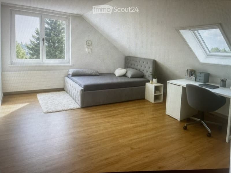 Cozy attic bedroom with wooden flooring, grey upholstered bed, nightstand, desk with chair, laptop, and small rug.