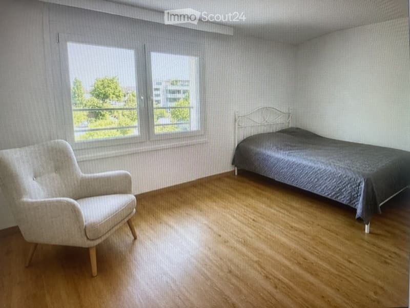 Bright bedroom with wooden flooring, large windows, a single bed with dark bedding, and a light-colored armchair.