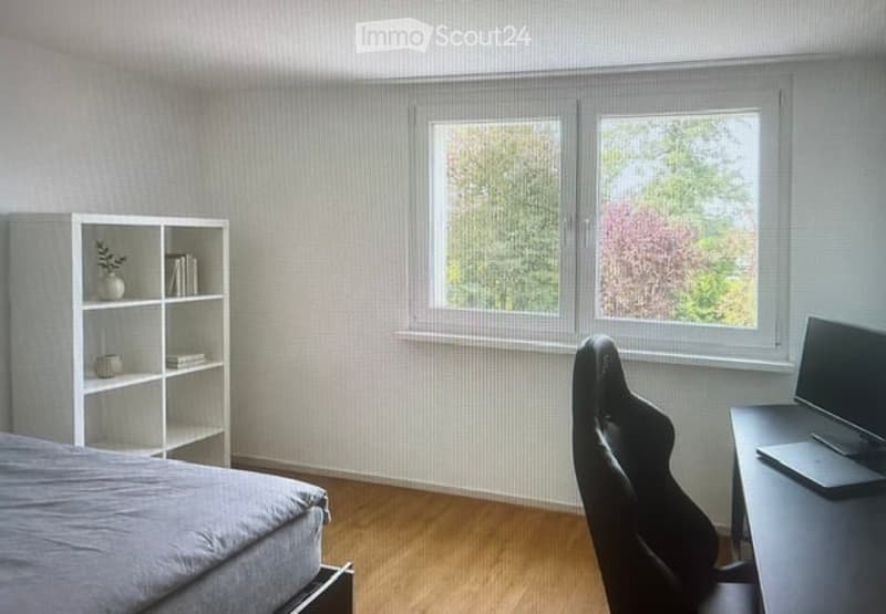 Modern bedroom with a large window, wooden floor, grey bed, bookshelf, desk with laptop, and a black gaming chair.