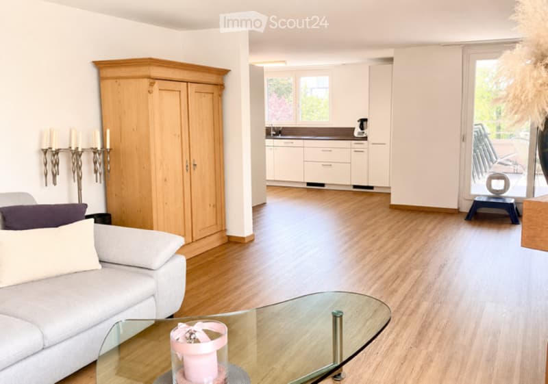 Bright, open-plan living and dining area with light wood flooring, white walls, a grey sofa, a glass coffee table, a wooden wardrobe, and a modern kitchen area with white cabinets and a sink.
