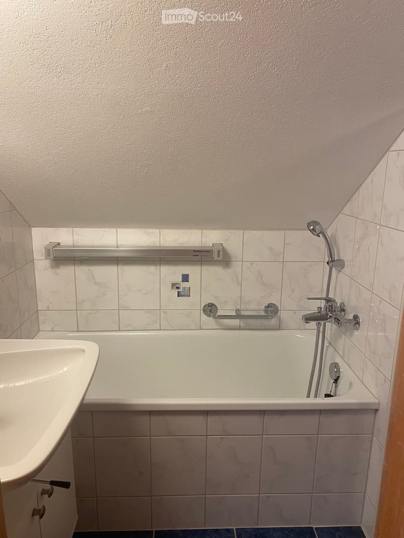 Bathroom with a bathtub, showerhead, faucet, and handrail. The walls are tiled with marble-like tiles, and the floor is covered with blue tiles.