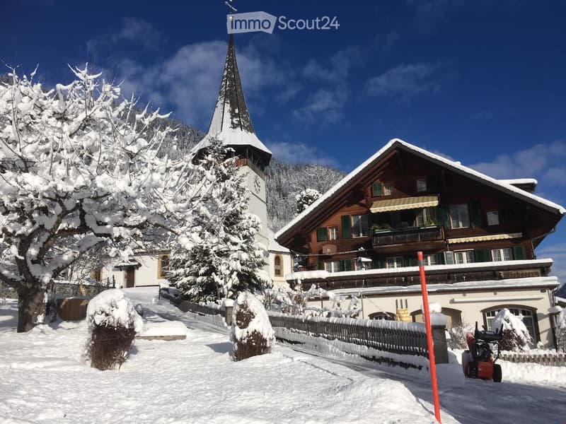 A traditional Swiss wooden house with green shutters and a balcony, surrounded by snow-covered trees and a church with a tall spire in the background.