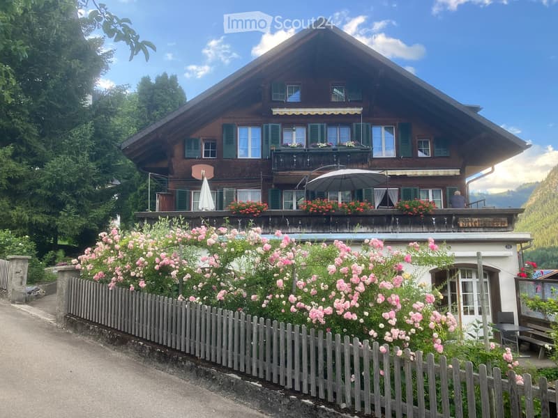 A two-story wooden house with green shutters, a balcony adorned with flowers, and a garden with pink roses and a wooden fence.