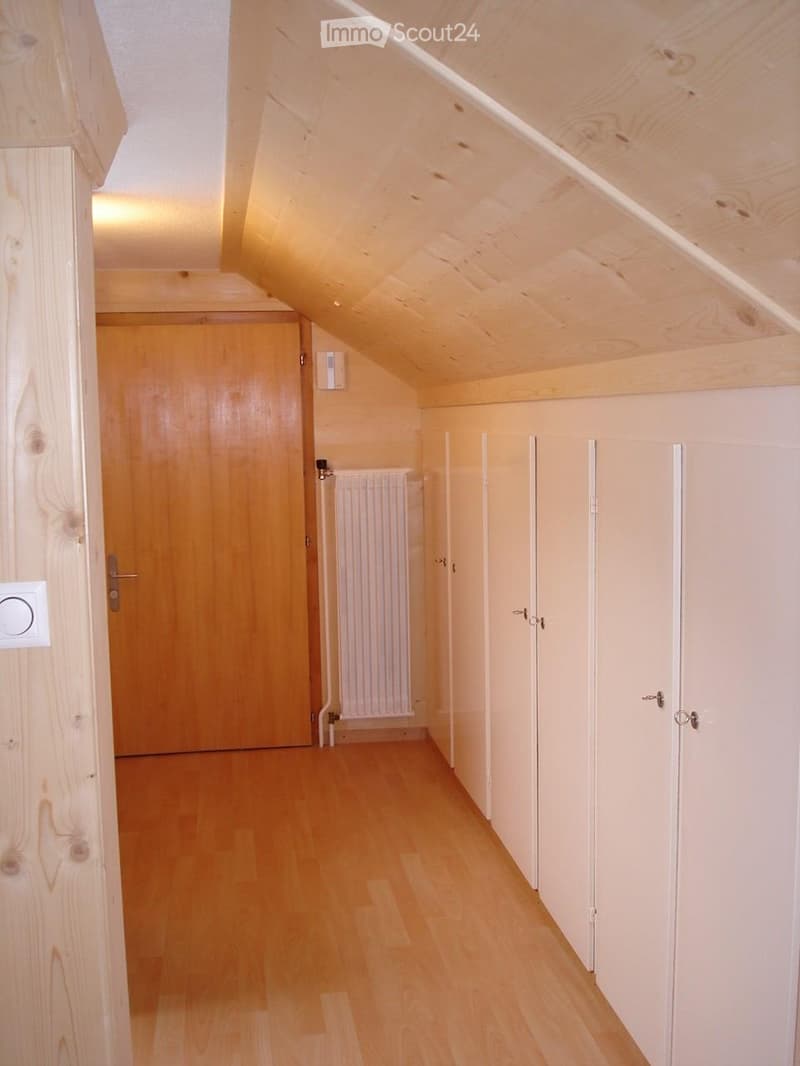 Bright hallway with wooden flooring, white walls, and several closed doors. The ceiling is slanted, typical of an attic space, and there is a radiator visible on one wall.