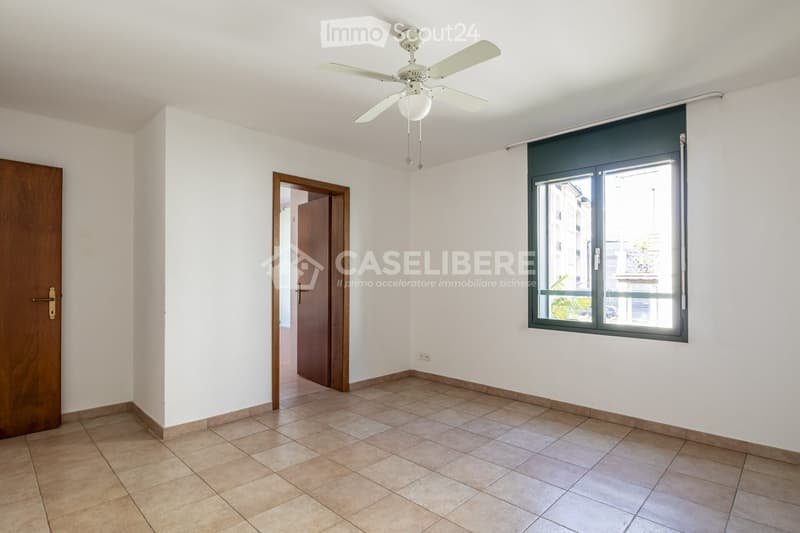 Empty room with white walls, tiled floor, wooden doors, and a ceiling fan with light fixture. Large window with blinds offers a view of the outside.