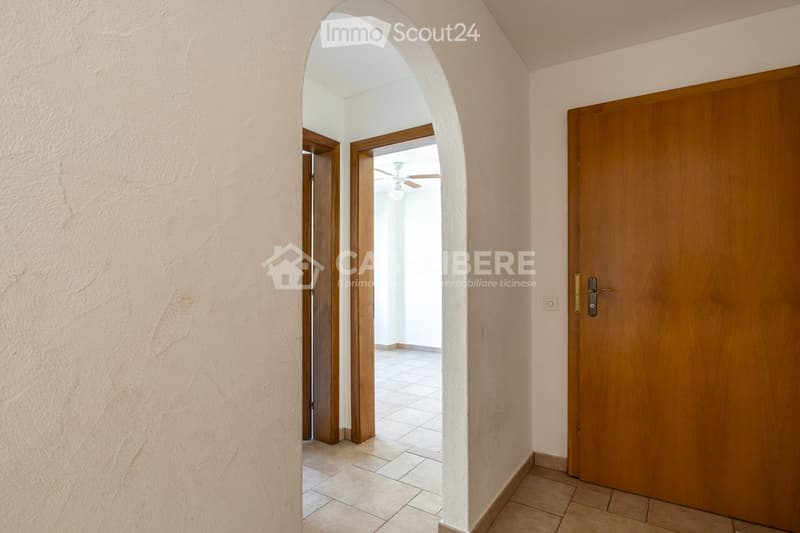 A well-lit hallway with beige tiled flooring, light-colored walls, and wooden doors. An arched opening leads to another room with a visible ceiling fan and light fixture.