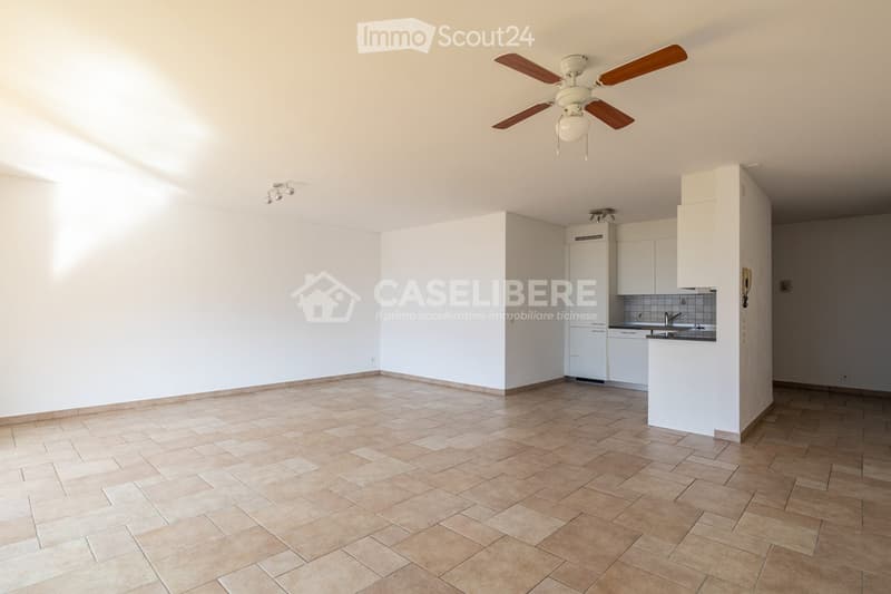 Empty room with tiled floor, white walls, and a small kitchen area with white cabinets and appliances.