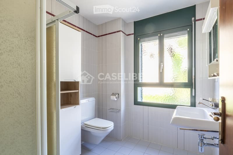 Modern bathroom with white tiles, a toilet, a sink with a chrome faucet, a mirror, and a wooden shelf cabinet.