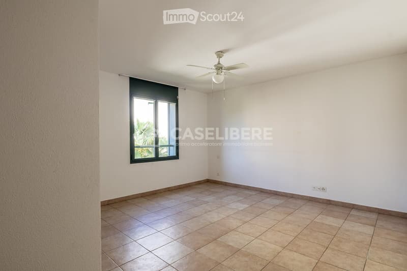Empty room with tiled floor, white walls, and a large window with blinds. A ceiling fan with a light fixture is mounted on the ceiling.