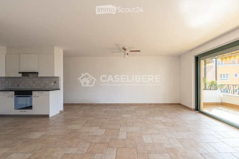 Open-plan kitchen and living area with tiled flooring, white cabinets, an oven, and a ceiling fan. Large sliding glass door leads to a balcony with outdoor plants.