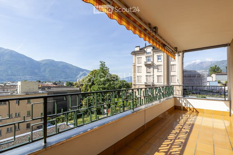Balcony with tiled floor, green metal railing, striped awning, overlooking cityscape and mountains.