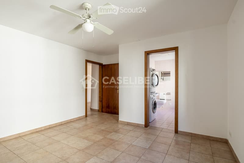Empty room with tiled floor, white walls, and a ceiling fan. A doorway leads to a bathroom with a toilet, washing machine, and sink.