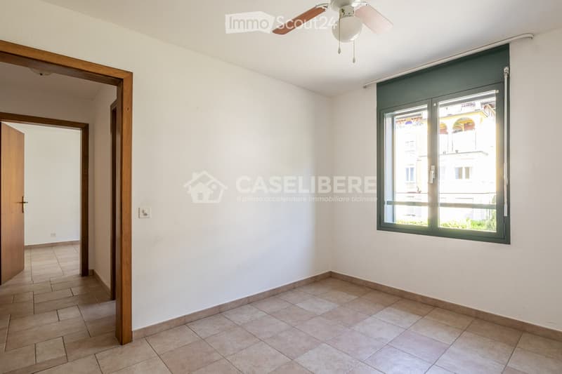 Empty room with tiled floor, white walls, wooden door frame, and a window with blinds. Ceiling fan visible.