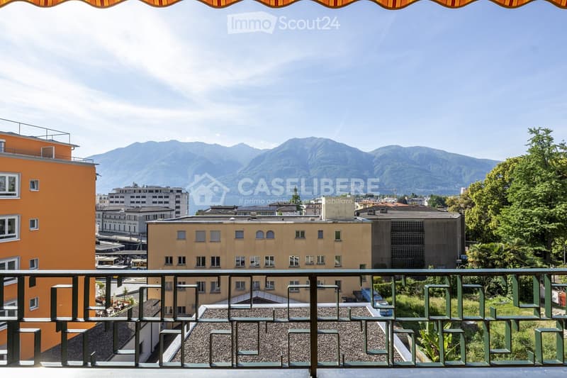 Balcony with metal railing overlooking a cityscape and distant mountains, with buildings and greenery visible below.