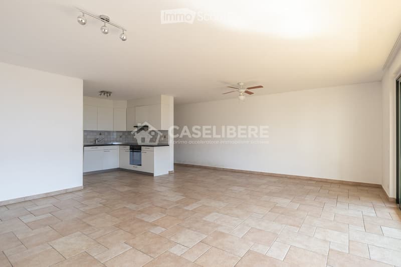 Empty room with tiled floor, white walls, and a small kitchen area with white cabinets and appliances.