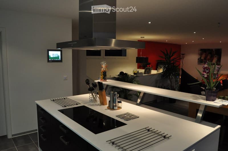modern kitchen with white countertops, black stove, stainless steel hood, black cabinets, white ceiling, red accent wall, TV mounted on wall, plants, and a dining table