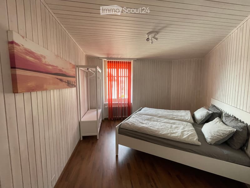 Cozy bedroom with wooden walls and ceiling, featuring a single bed with pillows, a small bench, and a painting on the wall.