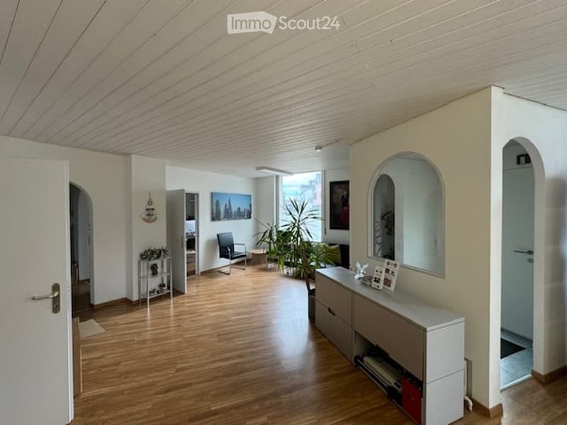 Bright, open living space with wooden flooring, white walls, and modern decor including a sofa, chair, small table, mirror, plant, and cabinet.