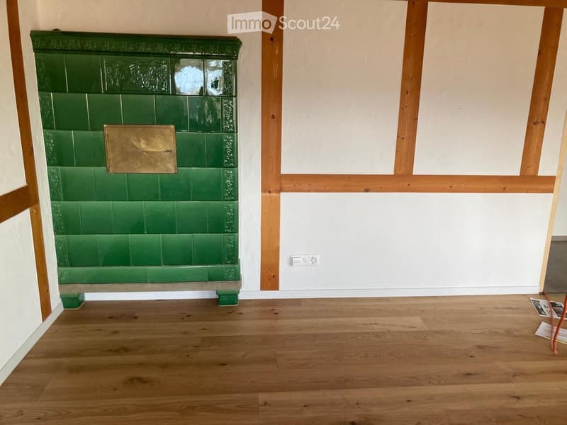 Empty room with wooden flooring, white walls, and a green tiled fireplace with a brass plaque.