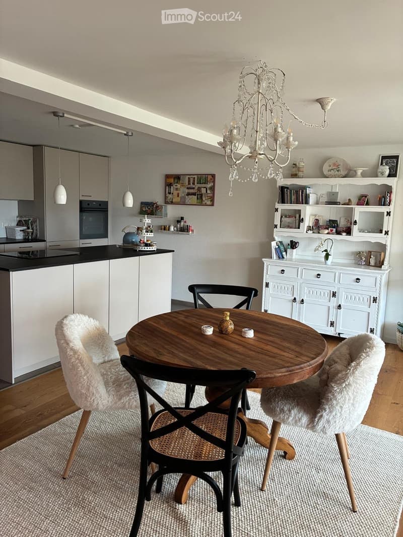 A modern dining area with a round wooden table, four chairs, a chandelier, and a kitchen area with white cabinets and an oven. The space includes a bookshelf and decorative items on the walls.