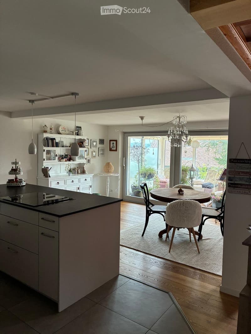 Modern kitchen with a central island, white cabinets, pendant lights, and a dining area with a round table and chairs. A large sliding glass door leads to a garden.