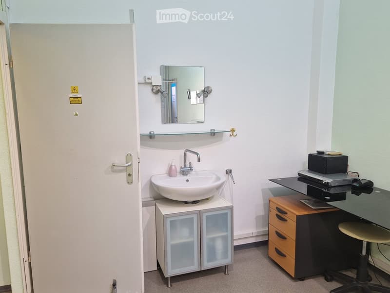 Modern bathroom with a white sink, mirror, glass shelf, and a small wooden cabinet. A desk with a chair is also present.