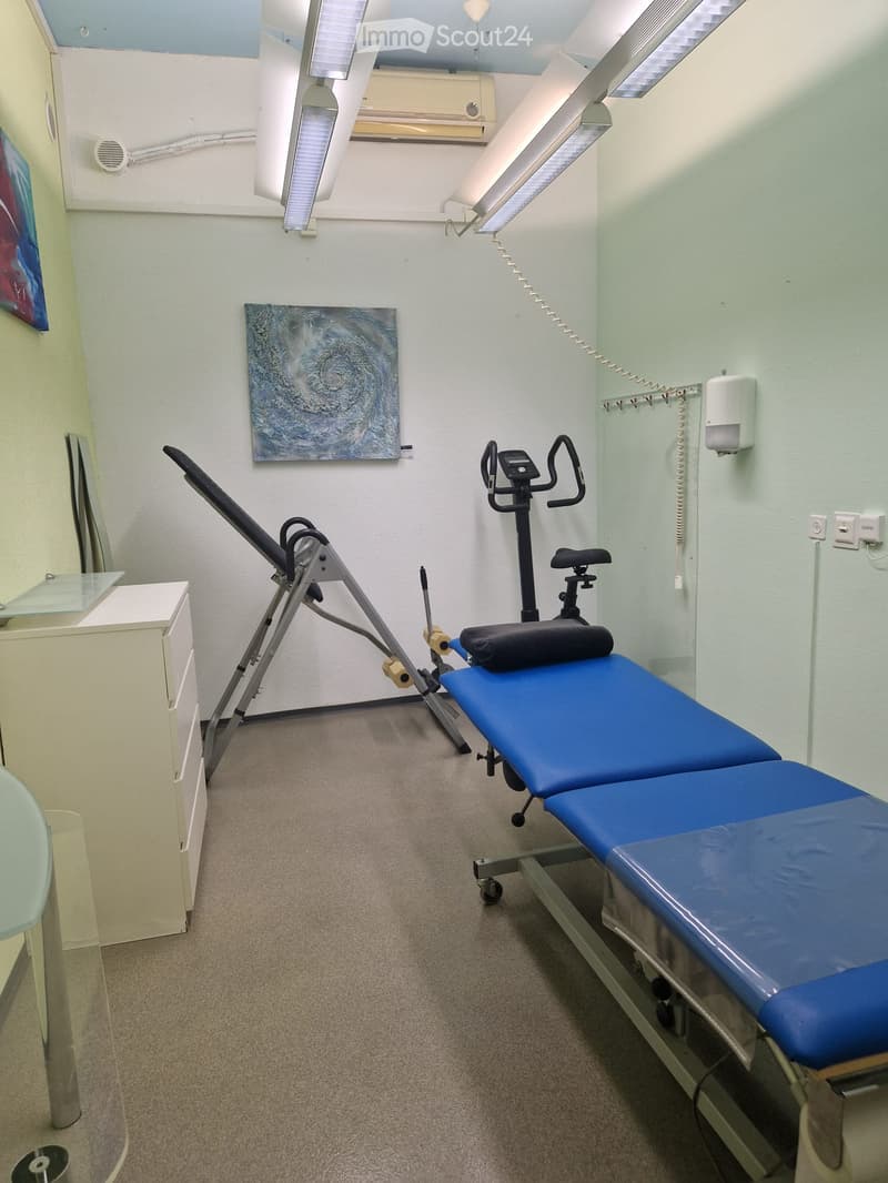 A clean, well-lit room with a treatment table, exercise machine, cabinet, and artwork on the walls.